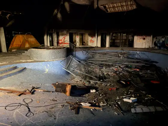 Drained blue-tiled swimming pool filled with collapsed fencing and debris beneath a damaged ceiling.