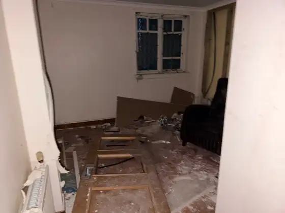 Small damaged hotel room containing loose boards, broken wall panels and a black chair beside the window.