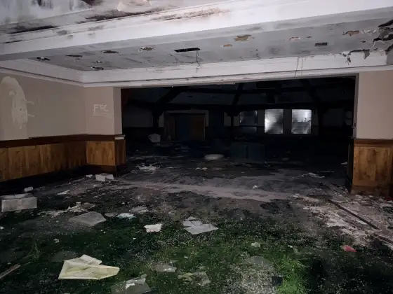 Wide opening into a dark wood-panelled function hall with boarded windows, ceiling damage and debris on patterned carpet.